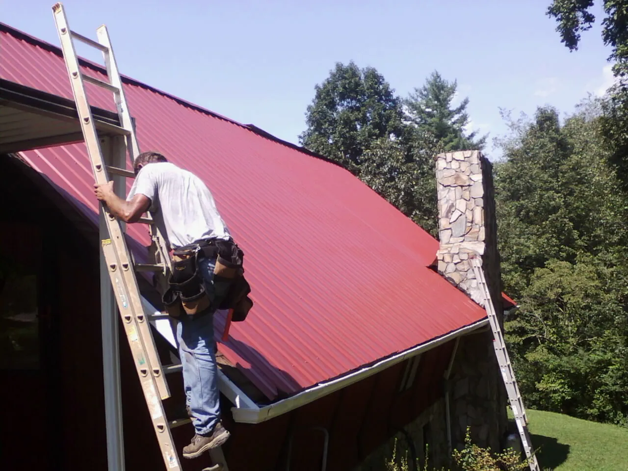 Roofer on a ladder beside a red metal roof and stone chimney — Soffit & Fascia Repair by Roofing Brothers in Dilworth